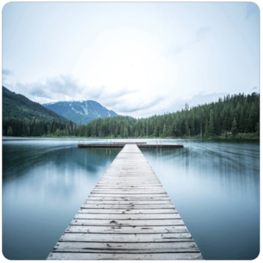A dock leading out into a lake, with a gray, cloudy sky and mountains in the background.